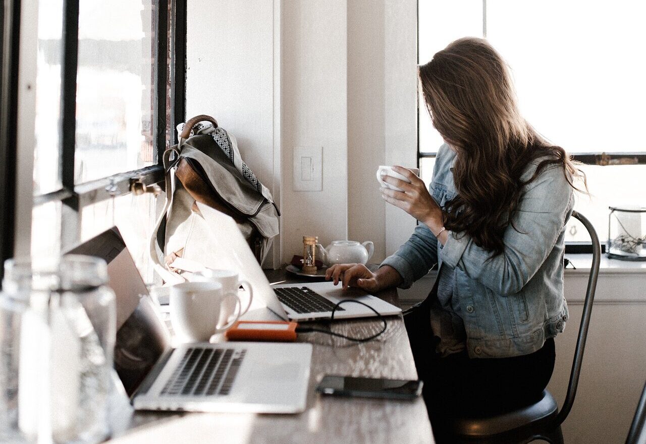 Kobieta trzyma kubek, siedzi przy biurku i pracuje przy laptopie