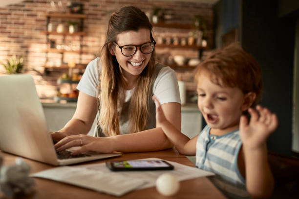 Kobieta w okularach pracje na komputerze, a obok niej bawi się dziecko
