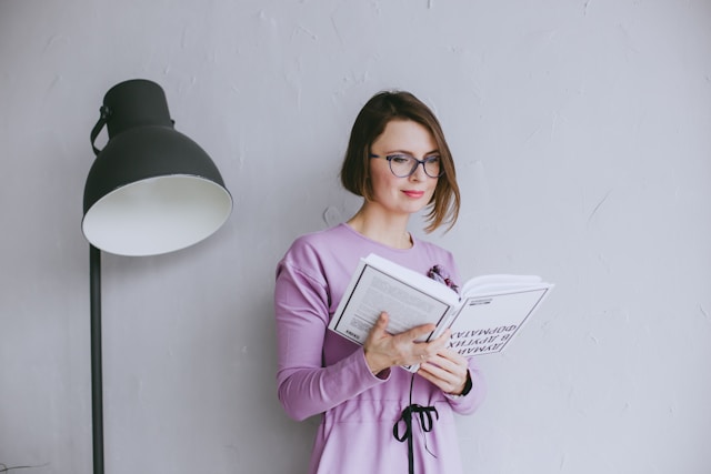 Kobieta we fioletowek bluzce stoi przy lapmie i czyta ksiażkę